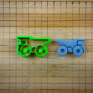 May include: Two plastic toy dump trucks, one green and one blue, on a wooden surface with a grid pattern.