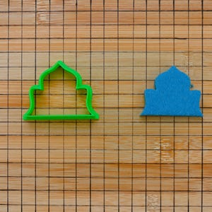 Può includere: Un tagliabiscotti in plastica verde e una forma in feltro blu, entrambi a forma di moschea, sono esposti su un tagliere di bambù. Il tagliabiscotti è di un verde brillante, mentre la forma in feltro è blu tinta unita.