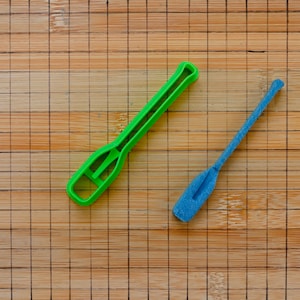 Peut inclure: Deux petites rames en plastique de couleurs vives reposent sur une planche à découper en bambou. L'une est vert vif et l'autre bleu vif. Les rames sont conçues avec une forme de pagaie et une poignée.
