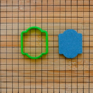 May include: Two cookie cutters on a wooden surface. The cookie cutter on the left is green and shaped like a rectangle with rounded corners. The cookie cutter on the right is blue and shaped like a rectangle with rounded corners.