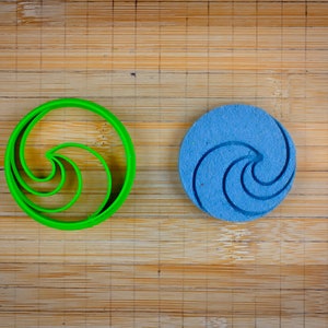 Peut inclure: Un emporte-pièce en plastique vert et un biscuit bleu en forme de spirale. Les deux sont sur une surface en bois.