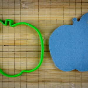 Peut inclure: Un emporte-pi&egrave;ce en forme de pomme en plastique vert et une pomme en feutre bleu sur une planche &agrave; d&eacute;couper en bambou.