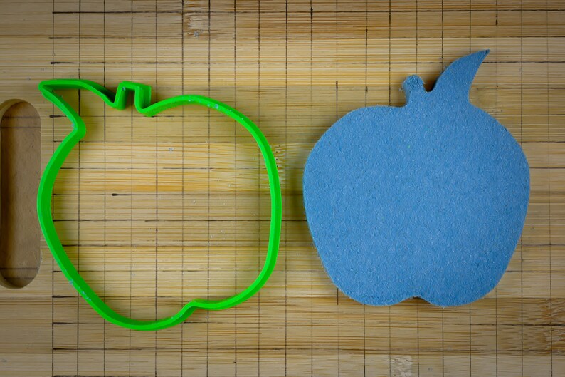 Peut inclure: Un emporte-pi&egrave;ce en forme de pomme en plastique vert et une pomme en feutre bleu sur une planche &agrave; d&eacute;couper en bambou.
