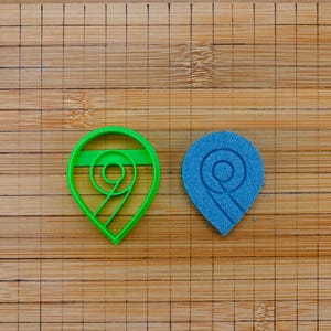 Pode incluir: Um cortador de biscoitos de plástico verde e um carimbo azul texturizado, ambos em forma de gota com um design em espiral, estão em uma tábua de corte de madeira. O cortador é verde brilhante, enquanto o carimbo é azul texturizado.