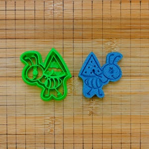 Può includere: Un tagliabiscotti in plastica verde brillante a forma di formica che trasporta una fetta di anguria, posizionato accanto a un biscotto blu testurizzato a forma di formica su un tagliere di legno con una griglia nera stampata.