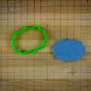 May include: A green plastic cookie cutter shaped like a banner or label, next to a blue felt banner or label.