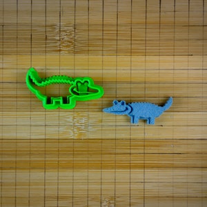 May include: Two alligator cookie cutters, one green and one blue, on a bamboo chopping board.