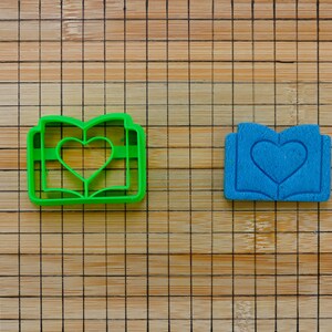 May include: Two cookie cutters shaped like an open book with a heart in the center. One is bright green, the other is blue. The cutters are on a bamboo cutting board.