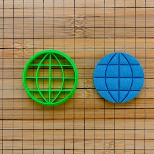 Op de afbeelding: Twee bolvormige koekjesvormen, een groene en een blauwe, op een houten oppervlak met een rasterpatroon.