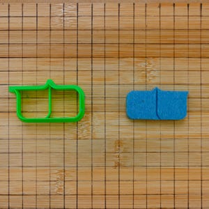 Puede incluir: Dos cortadores de galletas sobre una superficie de madera. Uno es verde brillante, el otro es azul. Ambos tienen forma rectangular con la parte superior curvada, diseñados para cortar masa o fondant.