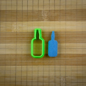 May include: Two green and blue plastic cookie cutters shaped like bottles. The cutters are on a wooden surface.
