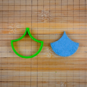 Puede incluir: Un cortador de galletas de plástico verde con forma de gota de agua, junto a una galleta de azúcar azul cortada en la misma forma.