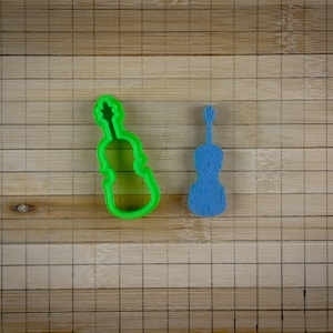 May include: A green plastic cookie cutter and a blue plastic cookie cutter, both shaped like a violin. The cutters are on a wooden surface.