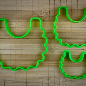 K&ouml;nnte beinhalten: Drei gr&uuml;ne Plastik-Ausstecher in Form einer L&auml;tzchen, ein gro&szlig;er und zwei kleinere. Die Ausstecher befinden sich auf einer Holzoberfl&auml;che.