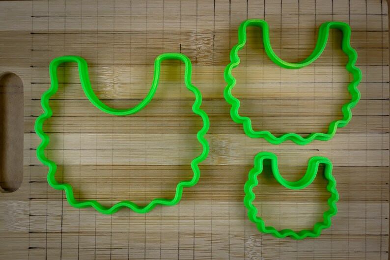 K&ouml;nnte beinhalten: Drei gr&uuml;ne Plastik-Ausstecher in Form einer L&auml;tzchen, ein gro&szlig;er und zwei kleinere. Die Ausstecher befinden sich auf einer Holzoberfl&auml;che.