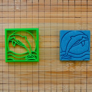 Könnte beinhalten: Ein grüner Delfin-Ausstecher aus Kunststoff und ein blauer Delfin-Stempel. Der Ausstecher ist ein quadratischer Rahmen mit einem Delfin, der über Wellen springt. Der Stempel hat das gleiche Delfin-Design.