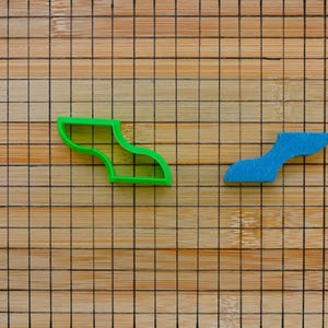 May include: Two cookie cutters on a wooden cutting board. The cutter on the left is green and shaped like a curved line. The cutter on the right is blue and shaped like a curved line.