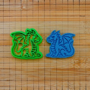 Peut inclure: Un emporte-pièce en plastique vert vif en forme de dragon placé à côté d'une empreinte de biscuit en forme de dragon bleu. Les deux objets présentent le contour détaillé d'un dragon avec des ailes, une longue queue et des écailles.