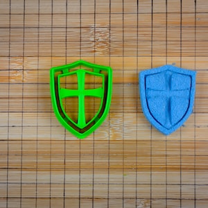 May include: Two cookie cutters in the shape of a shield with a cross inside. The shield on the left is green and the shield on the right is blue.