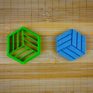 May include: Two hexagonal cookie cutters, one green and one blue, on a wooden surface. The green cutter has a geometric design and the blue cutter has a textured surface.