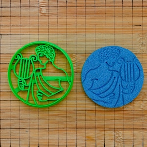 May include: A circular green plastic cookie cutter featuring an embossed design of a figure holding a lyre, positioned next to a matching blue circular cookie with the same embossed design of a figure holding a lyre, both on a grid surface.