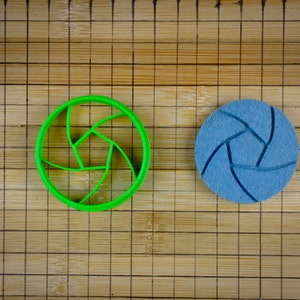 May include: Two round, 3D printed objects on a bamboo mat. The object on the left is green and the object on the right is blue. Both objects have a design that resembles a camera lens.