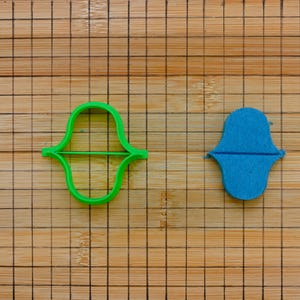 May include: Two cookie cutters in the shape of a stylized Hamsa hand. One is bright green plastic, and the other is blue felt. They are placed on a wooden cutting board with a grid pattern.