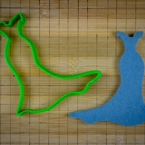 May include: A green plastic cookie cutter shaped like a dress, and a blue felt cutout of a dress on a wooden cutting board.