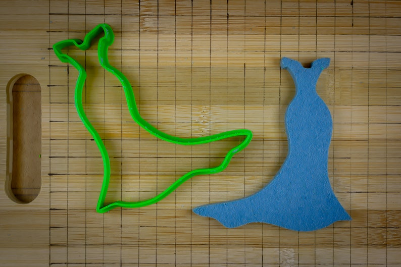 May include: A green plastic cookie cutter shaped like a dress, and a blue felt cutout of a dress on a wooden cutting board.