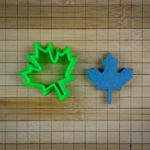 May include: A green plastic maple leaf cookie cutter and a blue felt maple leaf on a bamboo cutting board.
