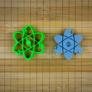May include: Two atom-shaped objects. One is a bright green plastic cookie cutter. The other is a light blue, star-shaped object with a similar atom design. Both are on a bamboo surface.