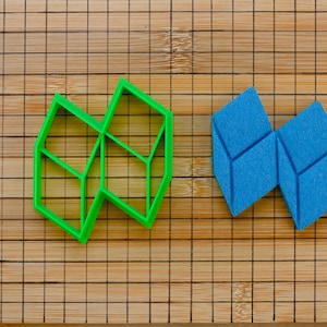 May include: A green geometric cookie cutter and two blue foam shapes sit on a bamboo cutting board. The cookie cutter has a complex, angular design. The blue shapes are also geometric, resembling connected cubes.