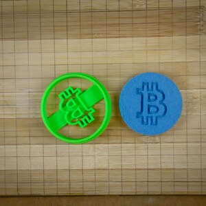 Peut inclure: Un emporte-pièce vert en forme de symbole Bitcoin avec une ligne diagonale qui le traverse, et un emporte-pièce bleu en forme de symbole Bitcoin.