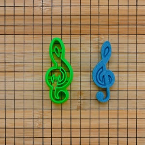 Puede incluir: Dos cortadores de galletas en forma de clave de sol, uno verde y otro azul, sobre una superficie de madera con un patrón de cuadrícula.