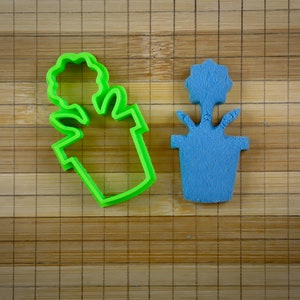 Può includere: Un tagliapasta in plastica verde a forma di pianta in vaso con un fiore in cima, accanto a una pianta in vaso in plastica blu con un fiore in cima.