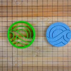 May include: A green plastic cookie cutter and a blue cookie with a wave design. The cookie cutter and cookie are on a bamboo cutting board.