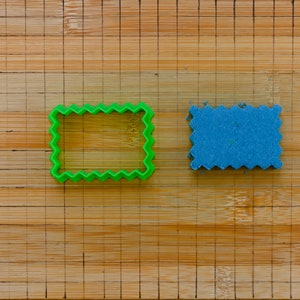 May include: A green plastic cookie cutter and a blue cookie on a wooden cutting board. Both the cookie cutter and the cookie are shaped like a rectangle with a wavy edge.