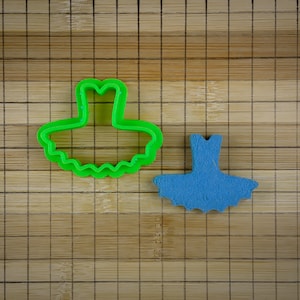 Può includere: Un tagliapasta di plastica verde a forma di abito da ballerina, e un abito da ballerina di fondant blu su un tagliere di bambù.