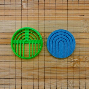 May include: A green plastic cookie cutter with a circular shape and a rainbow design sits next to a blue, circular sponge with a rainbow pattern. The items are on a bamboo cutting board.
