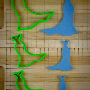May include: Three green plastic cookie cutters shaped like a dress with a long train, and three blue foam dress shapes on a wooden cutting board.