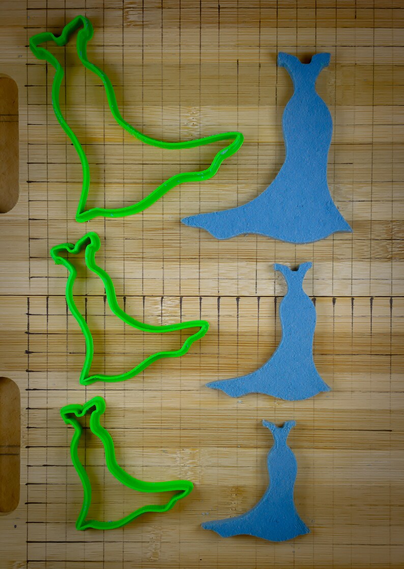 May include: Three green plastic cookie cutters shaped like a dress with a long train, and three blue foam dress shapes on a wooden cutting board.