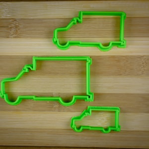 May include: Three green plastic cookie cutters shaped like delivery vans of different sizes on a wooden chopping board.