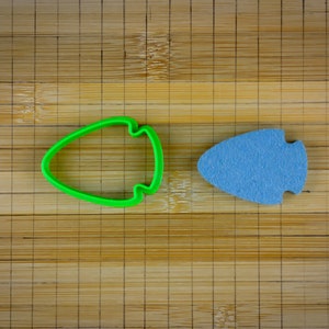 May include: A green plastic cookie cutter and a light blue foam cookie in the shape of a teardrop on a bamboo cutting board.
