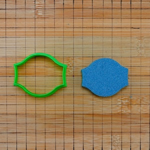 Può includere: Un tagliabiscotti in plastica verde e un oggetto blu strutturato sono su un tagliere di bambù. Il tagliabiscotti ha la forma di un'etichetta decorativa. L'oggetto blu ha la stessa forma.