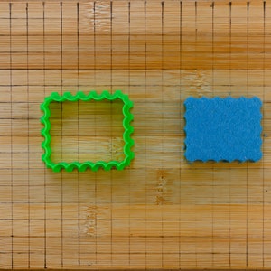 May include: A green plastic cookie cutter and a blue sponge, both with a scalloped edge, on a wooden cutting board with a grid pattern.