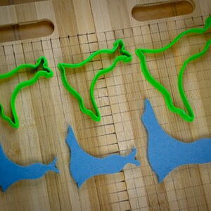 May include: Five cookie cutters on a wooden surface. Two are green and shaped like a stylized animal. Three are blue and shaped like a stylized animal.