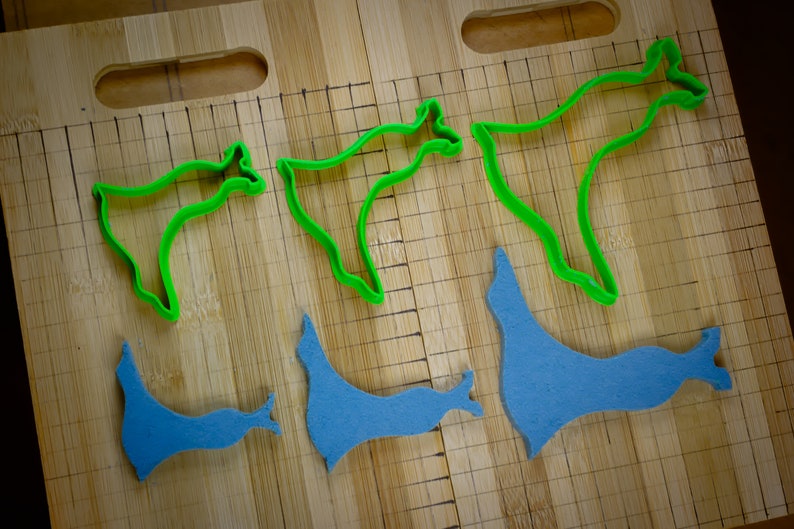 May include: Five cookie cutters on a wooden surface. Two are green and shaped like a stylized animal. Three are blue and shaped like a stylized animal.