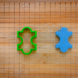 Könnte beinhalten: Zwei Ausstecher auf einem Holzbrett. Einer ist grün und hat die Form einer stilisierten Person. Der andere ist blau und hat die Form einer stilisierten Person.