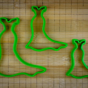 May include: Three green plastic cookie cutters shaped like dresses. The cutters are on a wooden cutting board.