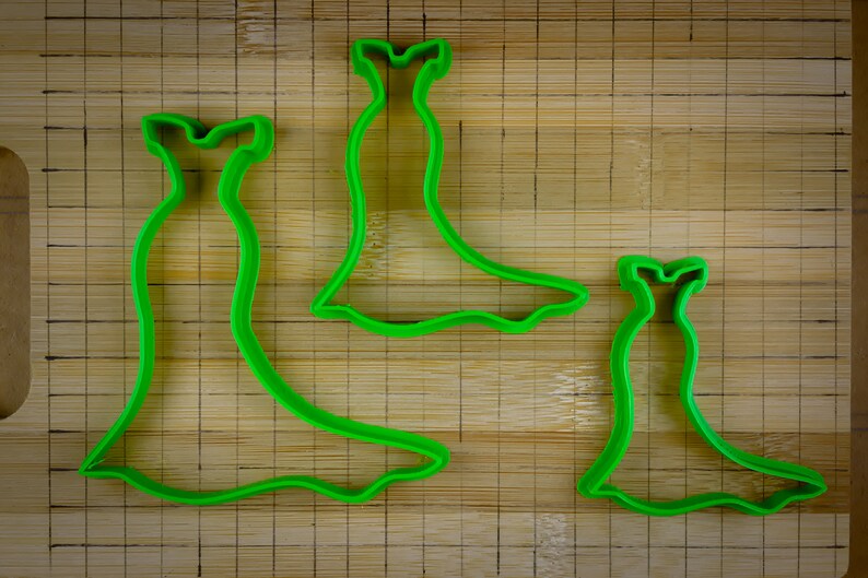 May include: Three green plastic cookie cutters shaped like dresses. The cutters are on a wooden cutting board.